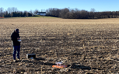 Staff member standing in a harvested corn fields looking at the drone controller. Drone is sitting on a orange circular disc in the field.