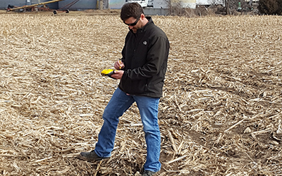 A staff member looking at a yellow GPS unit while standing over a small sinkhole in a harvested corn field.