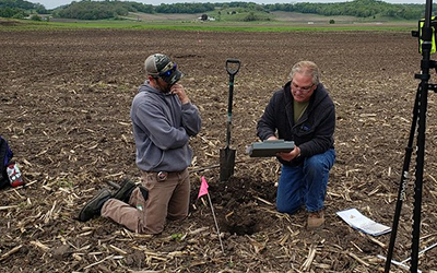 A staff member with a land owner. Both are kneeling over a small hole in the soil and looking at a clipboard.