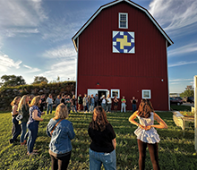 A circle of women with a large red barn in the background.