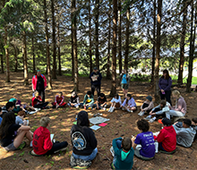 Youth sitting in a circle on the ground with pine trees in the background