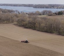 A tractor plowing a large field surrounded by trees, with a lake visible in the background.