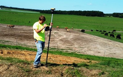 Man with a survey rod standing next to field with cows.